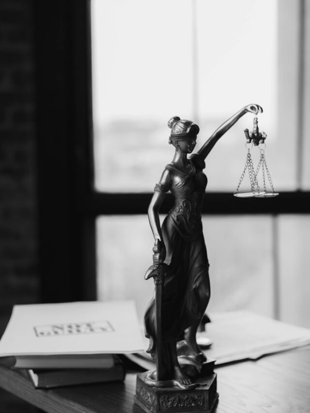 Grayscale photo of Lady Justice statue beside legal documents on a wooden table.